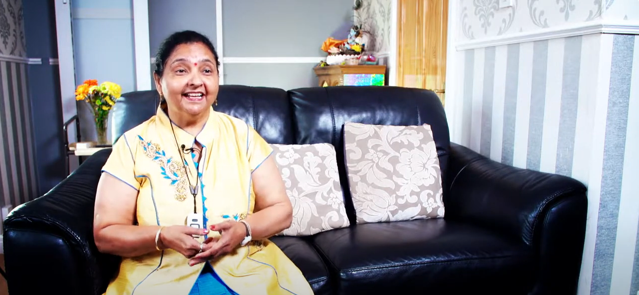 Mrs Parekh, smiling, sitting on her sofa while holding her lifeline pendant which is around her neck