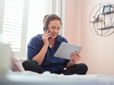 Lady sitting on herbed with her legs crossed, smiling holding her phone to her ear with her iPad in her other hand 